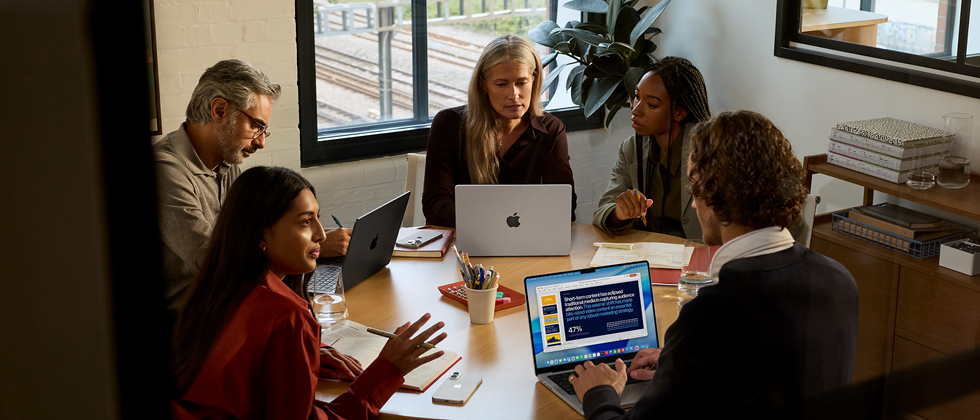 Un grupo de cinco empleados sentados en una mesa en una sala de reuniones, durante la reunión usan tres MacBook Pro.