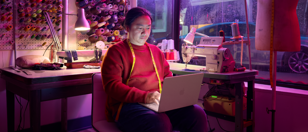 Una persona utiliza un MacBook Neo, color rosa nube, sin enchufar, sobre las rodillas, en un espacio de trabajo con una máquina de coser y materiales relacionados, como hilo y un maniquí