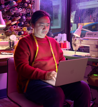 Una persona utiliza un MacBook Neo, color rosa nube, sin enchufar, sobre las rodillas, en un espacio de trabajo con una máquina de coser y materiales relacionados, como hilo y un maniquí