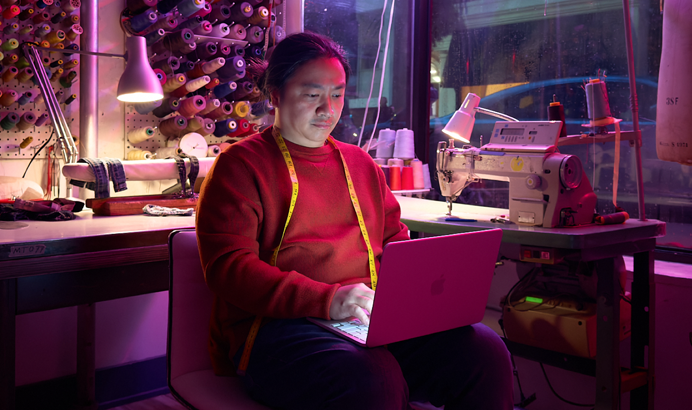 Una persona utiliza un MacBook Neo, color rosa nube, sin enchufar, sobre las rodillas, en un espacio de trabajo con una máquina de coser y materiales relacionados, como hilo y un maniquí